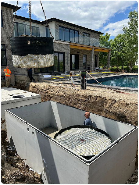 Installation of a residential septic system with a large filtration unit being lowered by crane into a concrete septic tank beside a luxury home with a swimming pool.